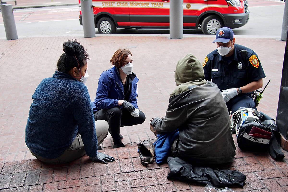 Three masked Street Crisis Response team members talk with a man seated on a step